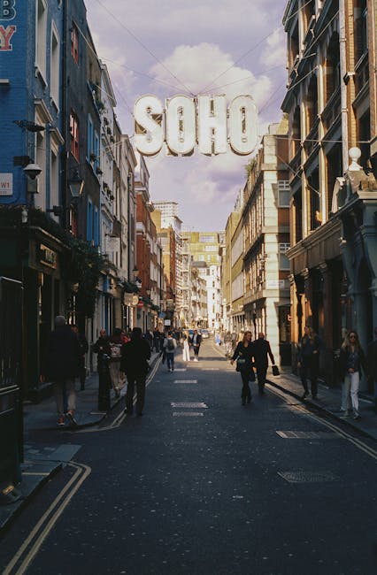 A bustling urban street scene at Carnaby Street in London, with a large illuminated sign spelling 'SOHO' hanging overhead across the narrow, pedestrianized road. The street is lined with multi-storey 