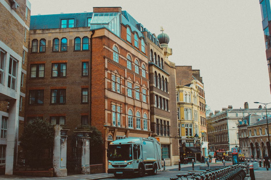 Two men are standing outside a building on a city street, engaged in a conversation during a home relocation process. One man is holding a black crate, and the other is pushing a wheeled cart. Nearby,