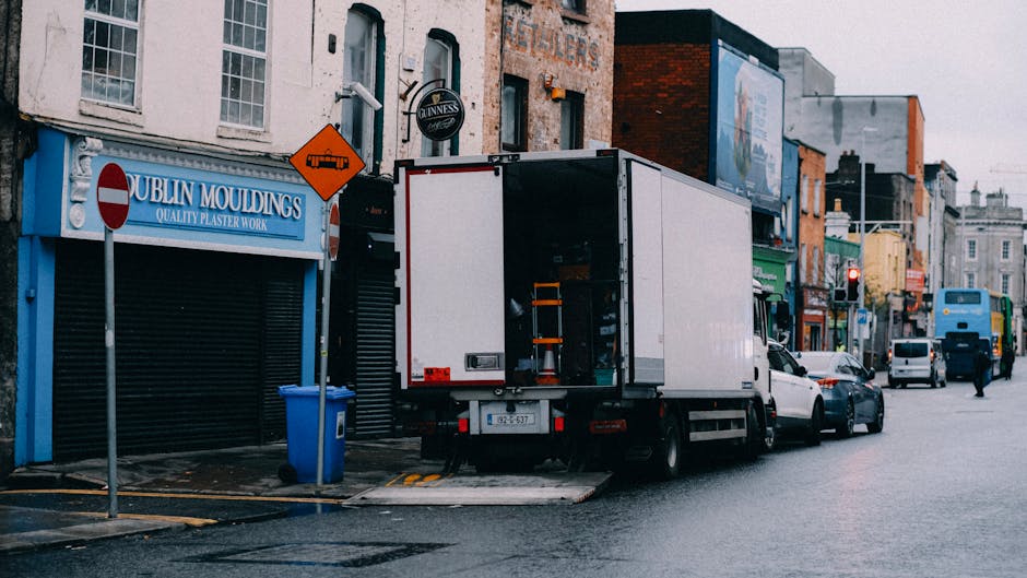 A white moving van with its rear doors open, positioned on a city street outside a shop called Dublin Mouldings, showing the loading process during a home relocation. Several cardboard boxes and wrapp