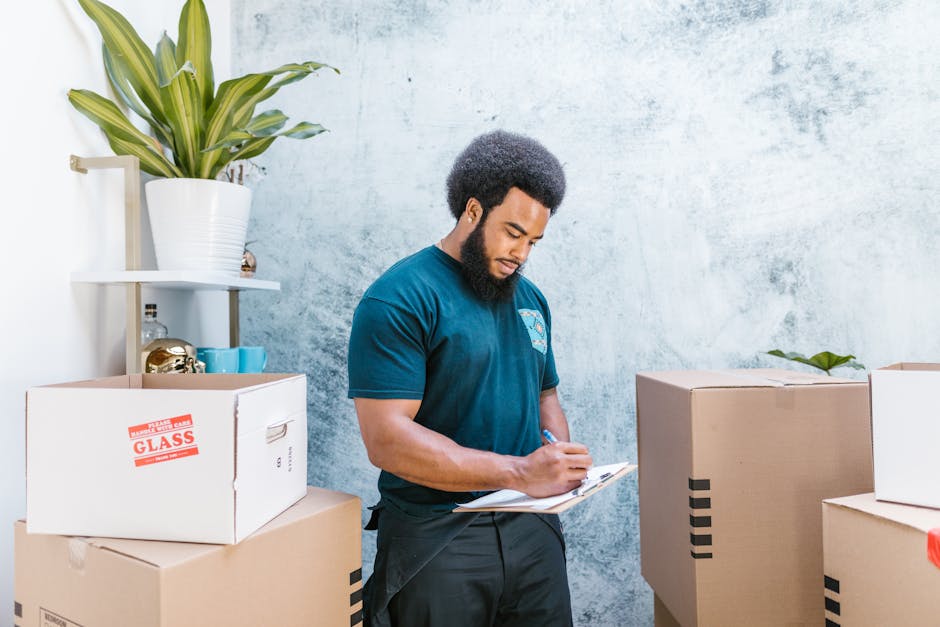 A man and a woman are engaged in a home relocation process inside a well-lit room with large windows. The man, standing and leaning over a cardboard box on a table, is holding a marker and appears to 